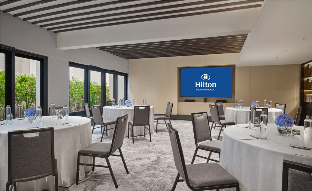 An elegantly arranged conference room with round tables set for a meeting, large screen displaying 'Hilton Singapore Orchard,' surrounded by windows with a view of greenery.