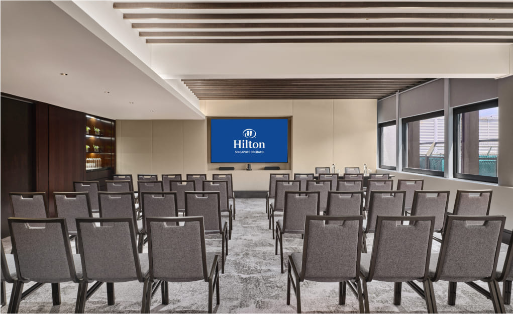 A conference room with rows of grey chairs facing a screen displaying the Hilton logo and 'Shanghai Gubei' underneath. Natural light filters through side windows.