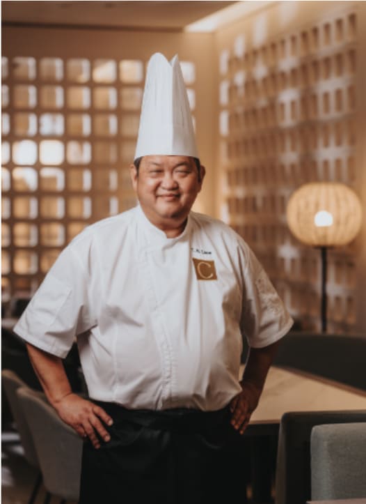 A chef in a white uniform and tall hat standing confidently with hands on hips in a well-lit, elegant dining room setting.