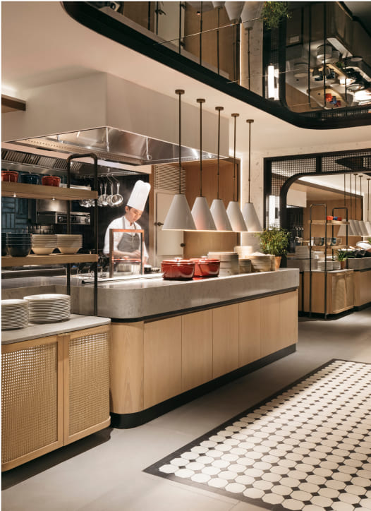 A chef is focused on cooking in a modern, well-lit commercial kitchen with wooden cabinetry, hanging utensils, and neatly arranged dishware.