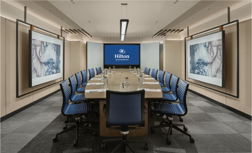 A modern boardroom with a long table, blue chairs, and screens on the wall displaying the Hilton logo.