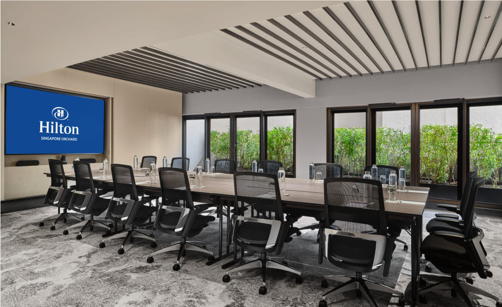 A modern conference room with a large display screen showing the Hilton logo and 'SINGAPORE ORCHARD.' Black office chairs surround a long table, alongside glassware, in daylight.