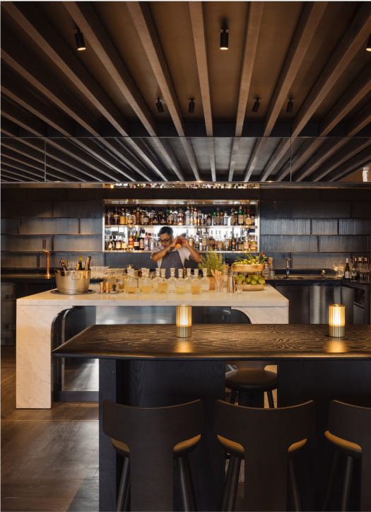 A bartender works behind a modern bar counter, surrounded by bottles and glasses, in a dimly lit, stylish bar with wooden ceiling details and dark furniture.