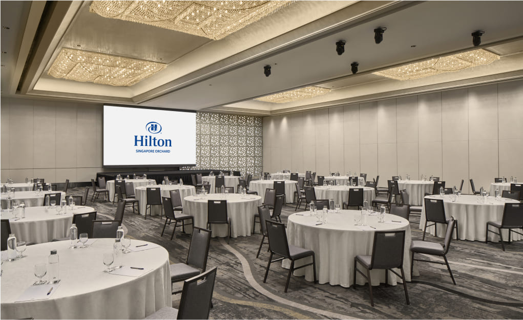 Round tables with white cloths are set for an event inside a spacious room with decorative lighting and a screen displaying 'Hilton Singapore Orchard'.