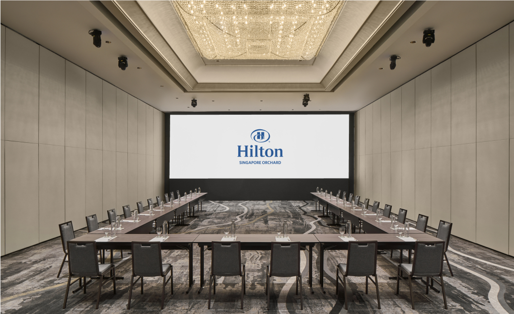 A long table with chairs and glasses is set for a meeting, with a large screen displaying 'Hilton Singapore Orchard' at the front of a well-lit conference room.