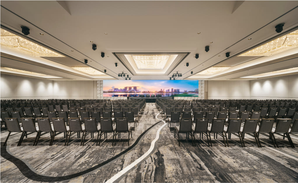 Rows of chairs are arranged to face a large screen displaying a cityscape within a spacious, well-lit conference hall.