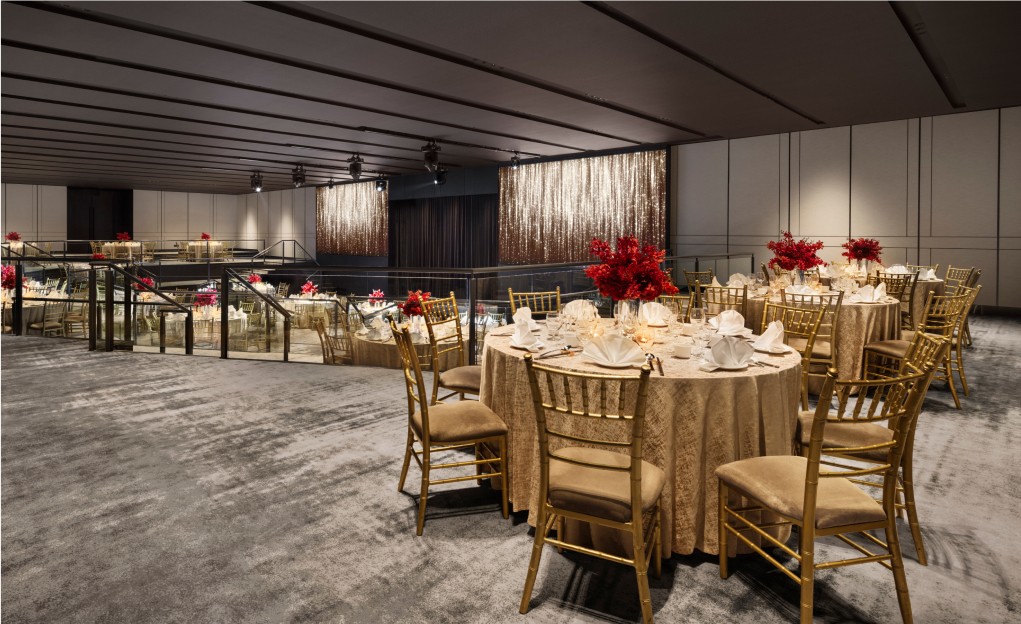 Elegant banquet hall set for an event with round tables, gold chairs, and red centerpiece flowers, under a subdued lighting and dark draped ceiling.
