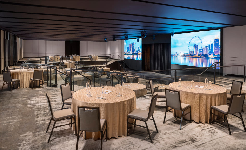 An event hall with round tables set for dining, overlooking a large screen displaying a city skyline, suggesting a formal gathering within a modern indoor setting.