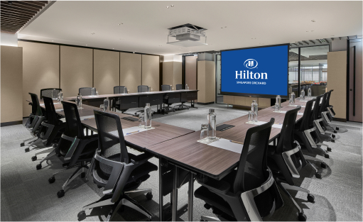 A modern conference room with a large table, chairs, and a screen displaying the Hilton logo, in a well-lit, indoor setting.