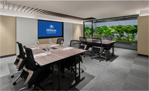 A modern conference room with a large table, chairs, and water bottles. A screen displays the Hilton logo. Large windows reveal lush greenery outside.