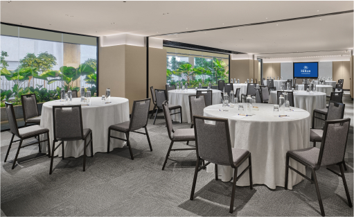 Round tables set with dining ware in a spacious conference room with large windows showing lush greenery outside.