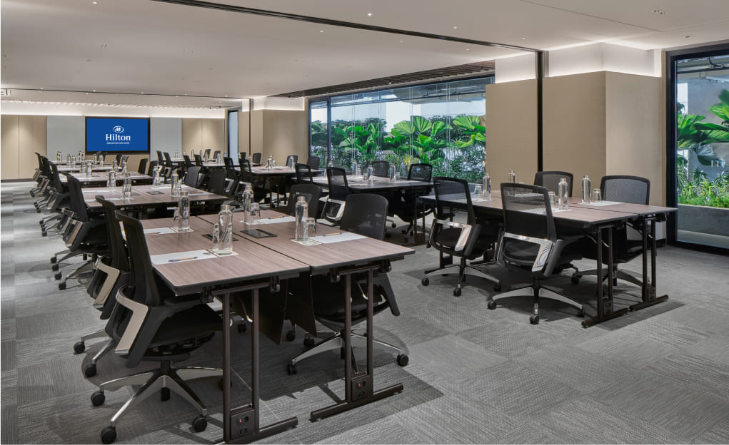 A spacious, modern conference room with arranged tables and chairs, a presentation screen displaying 'Hilton,' and large windows revealing lush greenery outside.