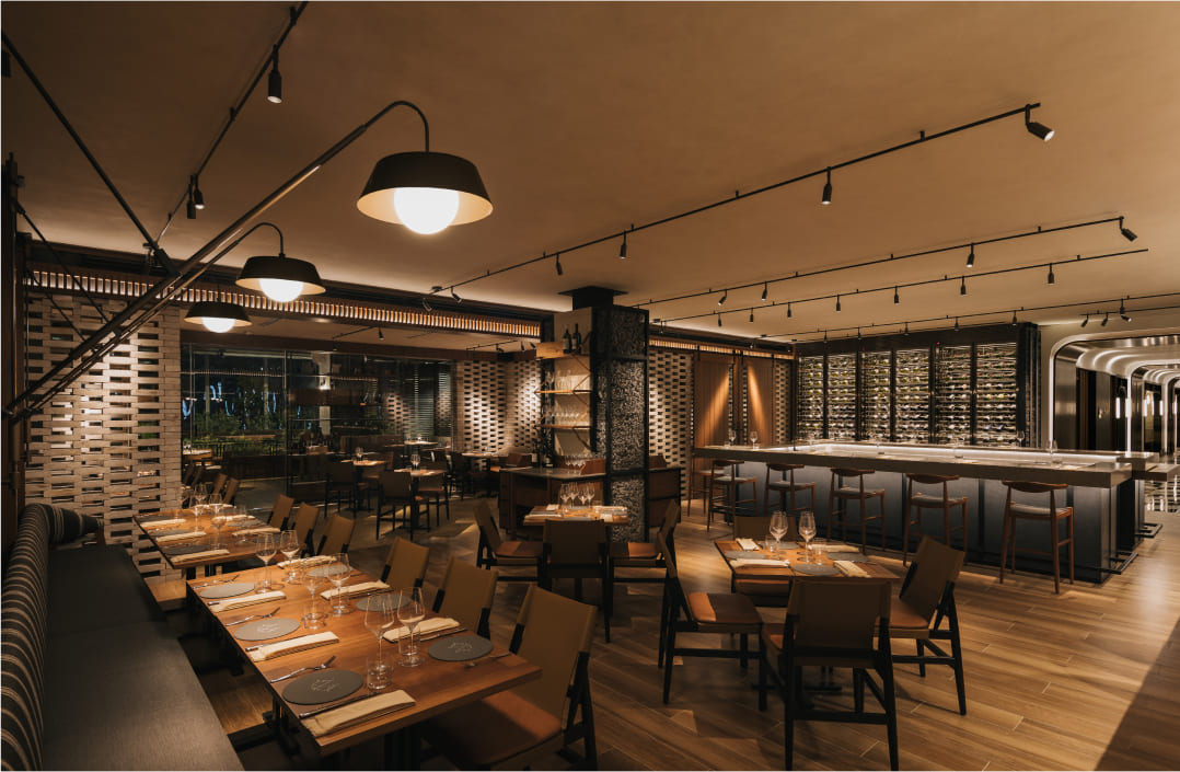Sophisticated restaurant interior with wooden tables set for dining, large industrial-style lamps overhead providing a warm ambiance, and wine bottles displayed to frame the cozy, modern space.