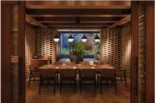 A warmly-lit dining room with a large table set for a meal, surrounded by chairs, in an elegant, modern setting with visible greenery outside.