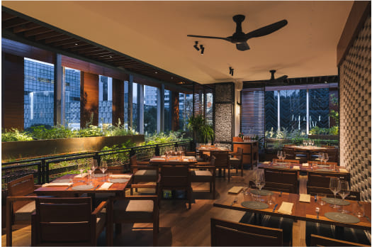 Elegant restaurant interior with set tables, warm lighting, and large windows looking out to city buildings at dusk. A ceiling fan is overhead, and plants line the windows.