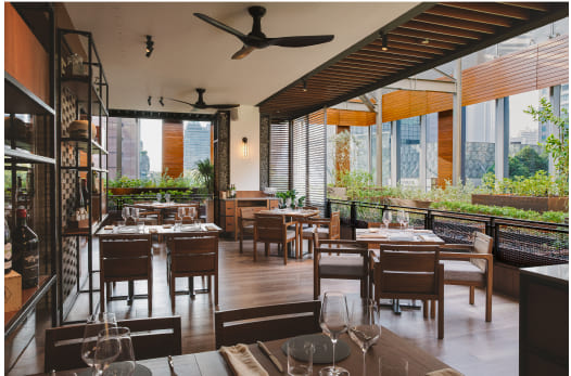 An elegant outdoor dining area with wooden furniture and glassware set on tables is surrounded by urban high-rise buildings and greenery.