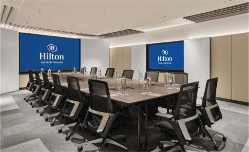 A rectangular conference table surrounded by office chairs in a modern meeting room with 'Hilton' logos on screens at one end.