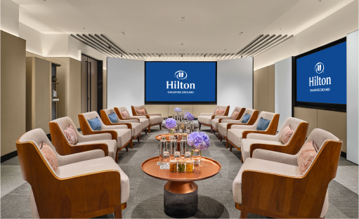 A modern meeting space with arranged chairs around a central table, facing two screens displaying the 'Hilton' logo. The context is an indoor corporate setting.