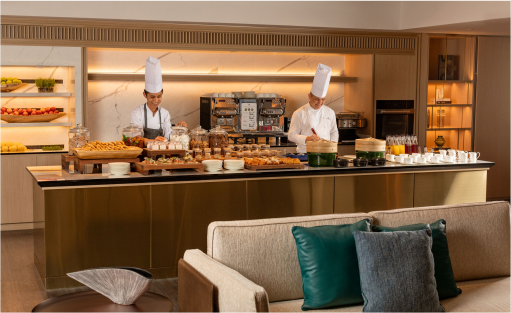 Two chefs prepare food behind a counter filled with various dishes in a hotel buffet, set against a modern and cozy dining area.