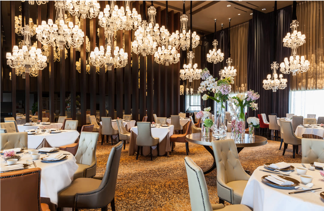 An opulent dining room featuring multiple crystal chandeliers, elegantly set tables with white linen, and a variety of upholstered chairs, all set upon a patterned carpet.