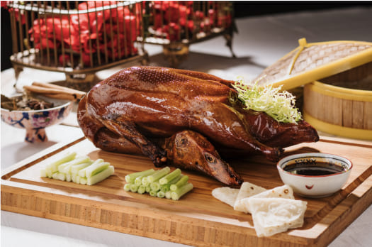 A roasted duck on a wooden board, accompanied by sliced cucumbers, spring onions, steamed buns, and a bowl of sauce, with a bamboo steamer in the background.
