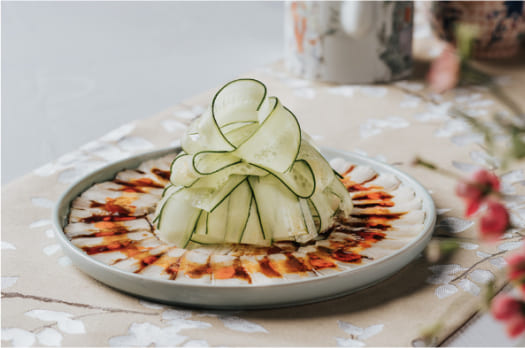 A decorative food arrangement resembling a flower made from thinly sliced cucumber, atop a mound of food on a plate with drizzled sauce, on a floral tablecloth.