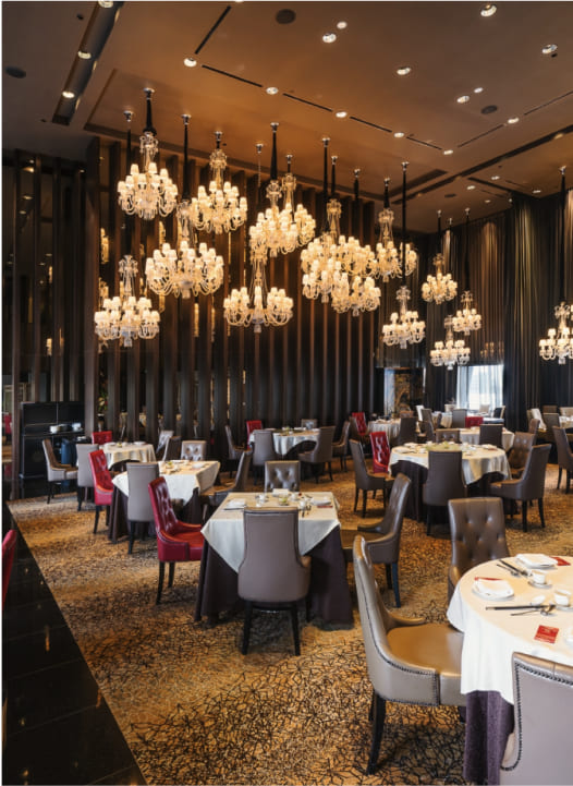 Elegant dining room with multiple chandeliers, set tables with red and white accents, surrounded by dark curtains and patterned flooring.