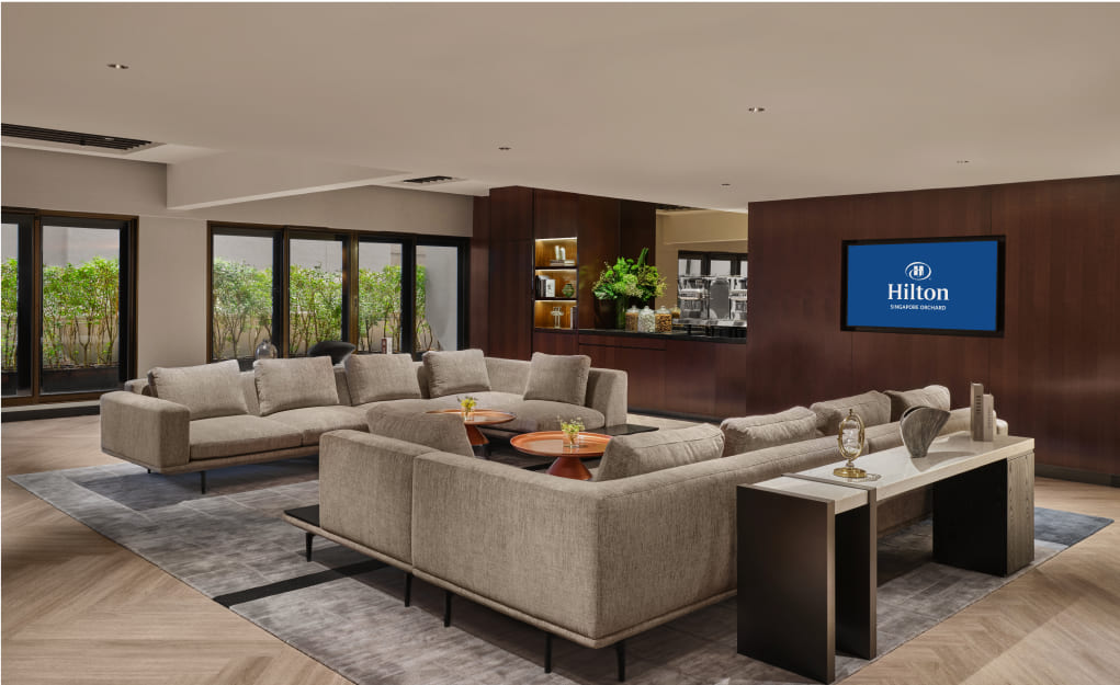 A luxurious hotel lounge with large beige sofas, a flat-screen displaying 'Hilton' on the wall, surrounded by windows revealing outdoor greenery.