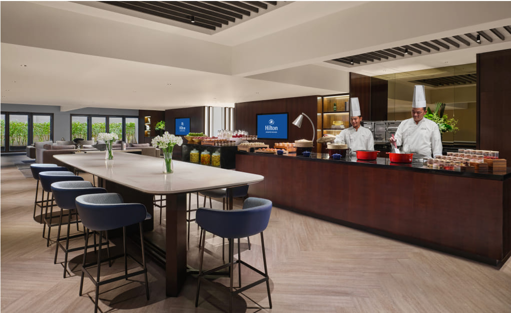 Two chefs prepare food behind a counter in a modern hotel kitchen with seating areas and large windows. The logo 'Hilton' is visible.
