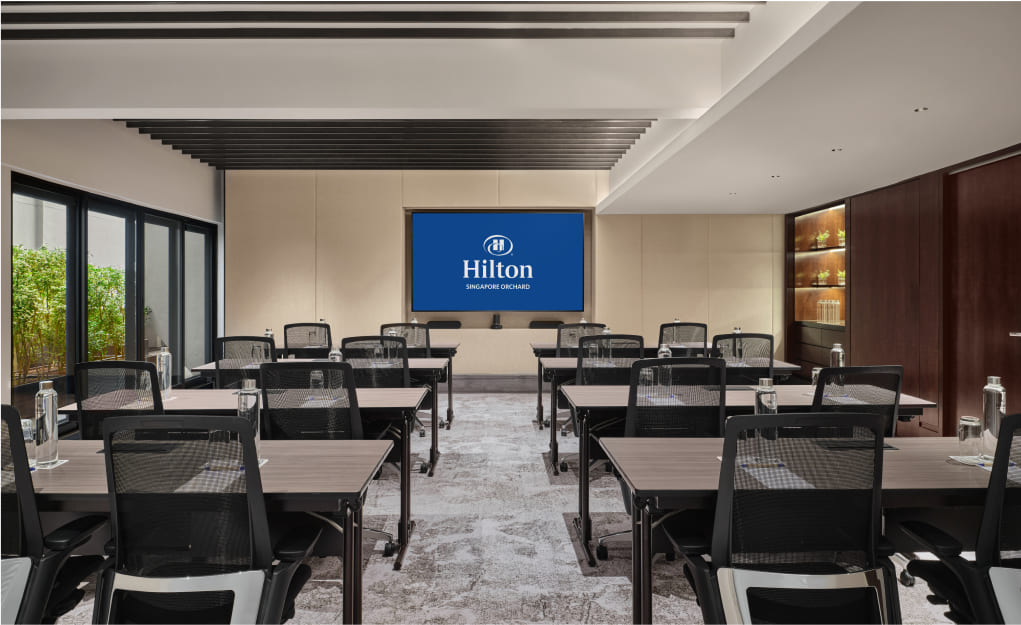 Conference room with arranged chairs and tables, large screen displaying 'Hilton' logo, ambient lighting, adjacent to glass windows showing greenery.