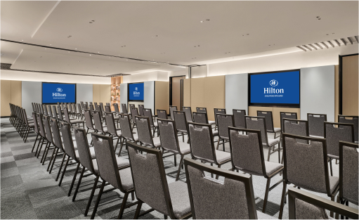 Rows of empty chairs face projection screens displaying the Hilton logo, set up in a well-lit conference room.