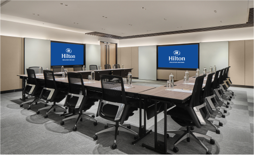 A rectangular meeting table surrounded by chairs in a room with screens displaying the Hilton logo, suggesting a business or hotel conference setting.